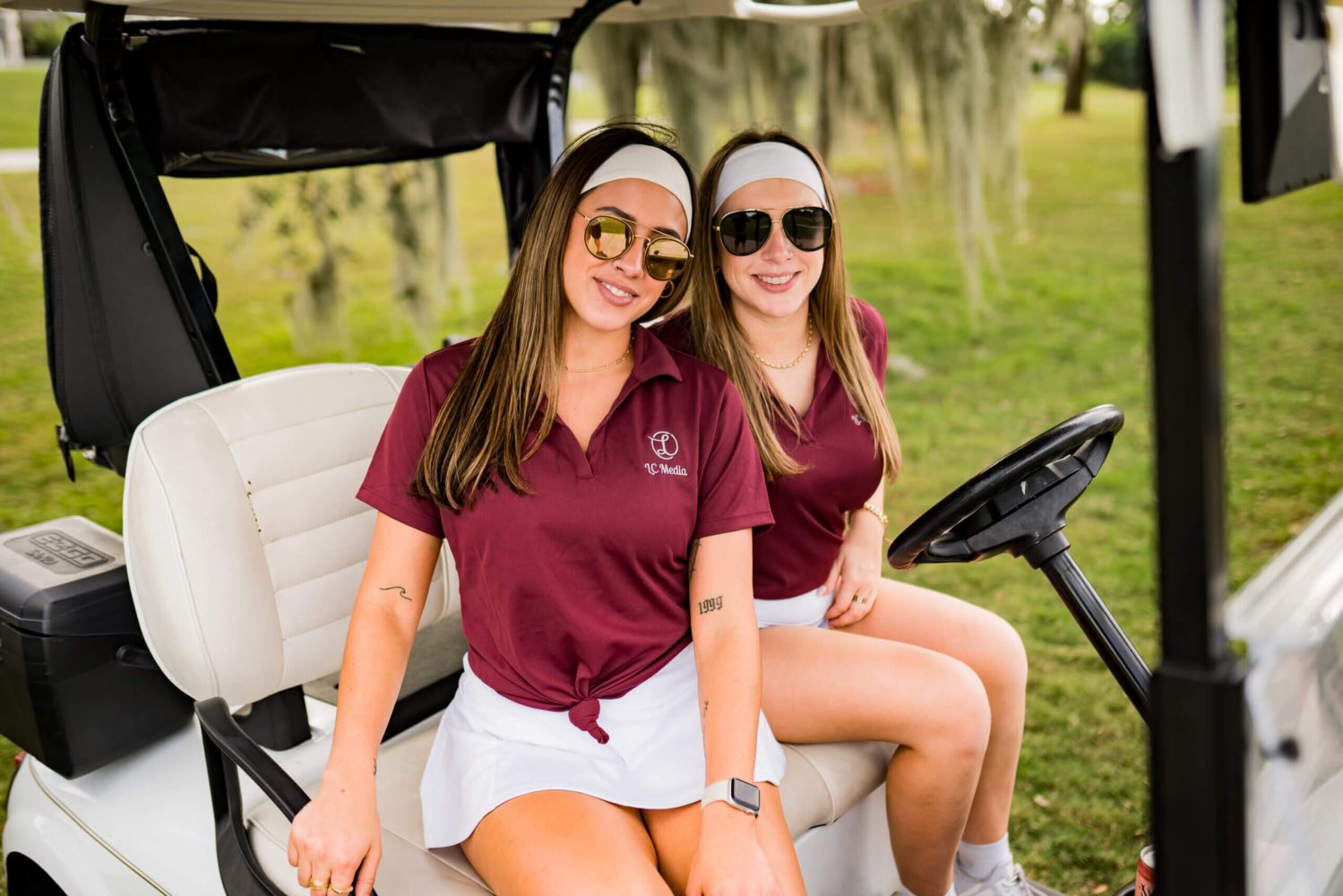 Two women sitting on a golf cart wearing maroon shirts.