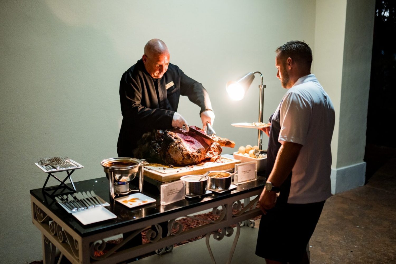 Two men standing around a table with food on it.