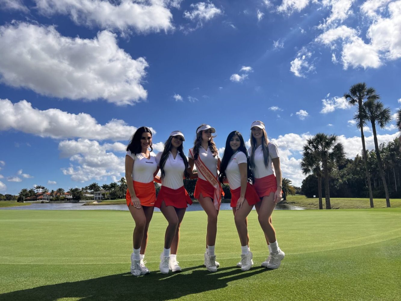 Four women in white shirts and red shorts standing on a green.