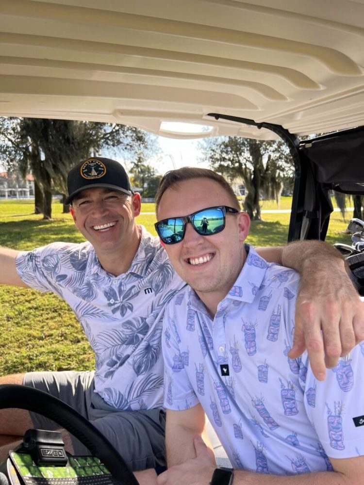 Two men in matching shirts and hats are sitting on a golf cart.